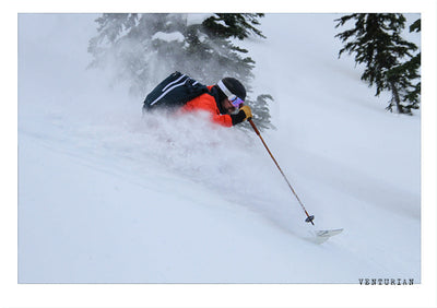 a venturian skiier charging through powder snow on skis in a red jacket