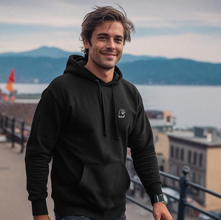 Man wearing a black hoodie with a logo, standing on a bridge with mountains and water in the background.