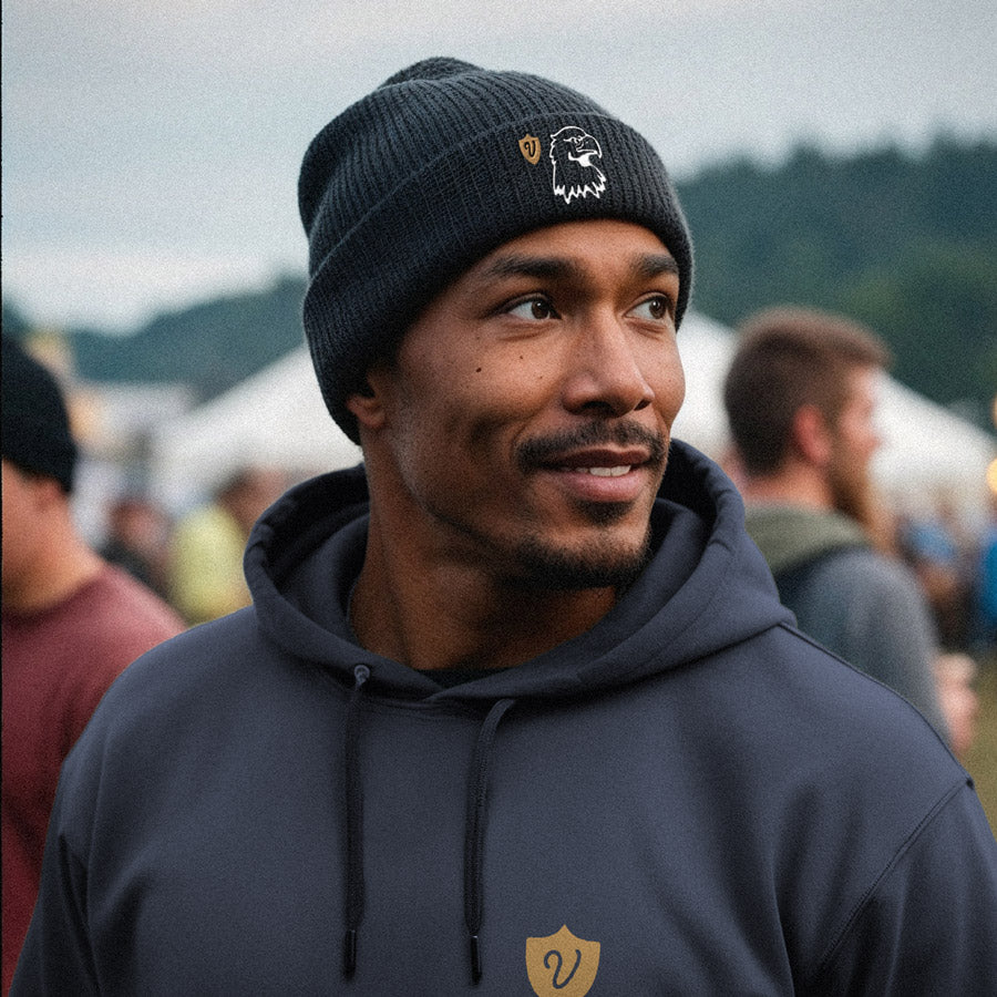Man wearing a black beanie and hoodie with a logo, standing outdoors with blurred people and trees in the background.