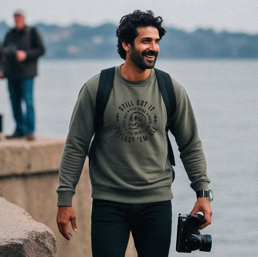 Man walking by a body of water holding a camera, wearing a green sweatshirt with skull.