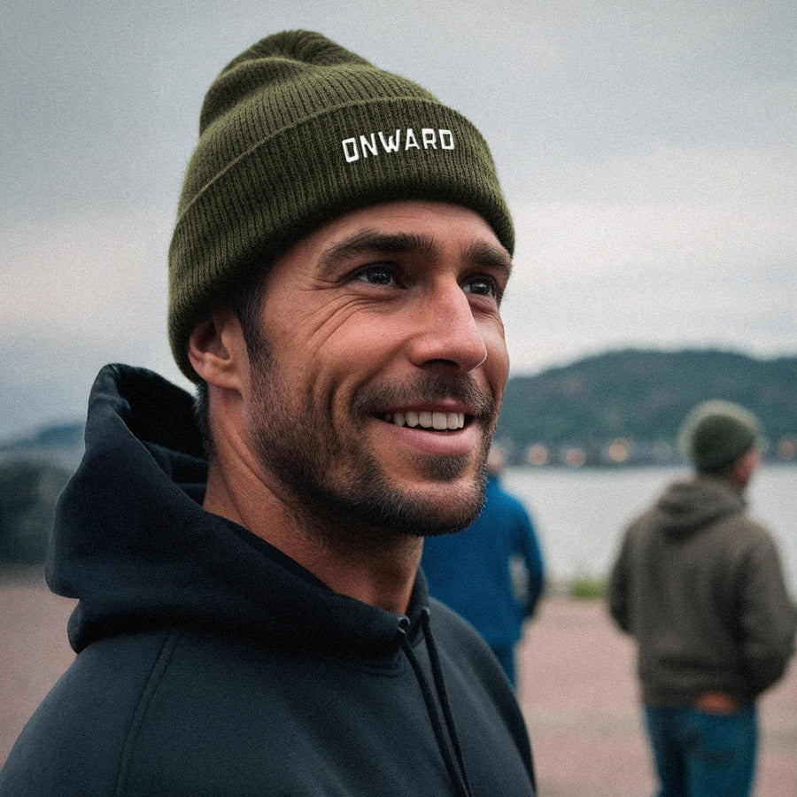 Man wearing a green 'ONWARD' beanie and black hoodie by a lake with mountains in the background.