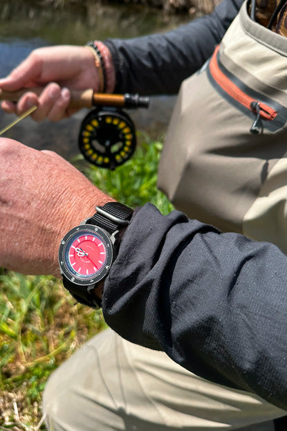 Person holding a flyfishing rod with a watch on their wrist, outdoors near water