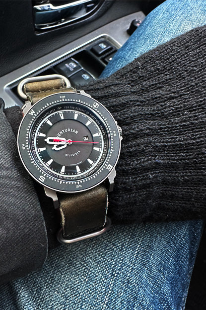 Watch with a brown leather strap on a person's wrist, sitting in a car.