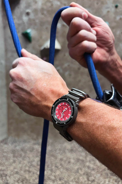 Person climbing with a watch on wrist, focusing on the watch's red face.