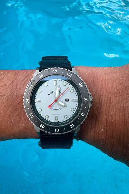Black wristwatch with a white face on a person's wrist by a pool.