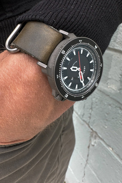 Watch with a black strap worn on a wrist against a gray wall.