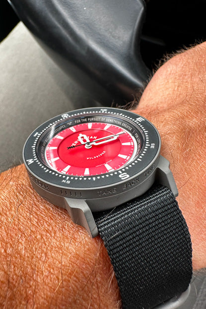 Watch with a red face and black strap on a wrist, with a blurred background.