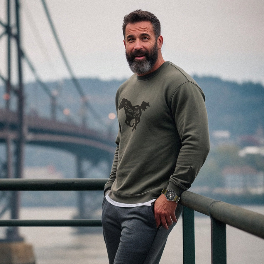 Man wearing a green sweatshirt with a horse graphic, standing by a railing with a bridge and water in the background.